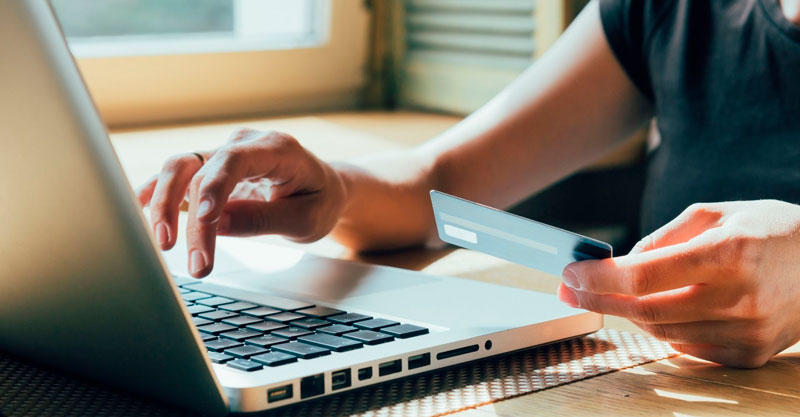 Mão de um homem, com camisa cinza, fazendo compra no seu laptop usando um cartão de crédito