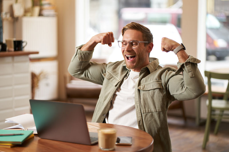 Retrato de jovem feliz com óculos comemorando sentado em um café assistindo a um jogo de esportes no laptop